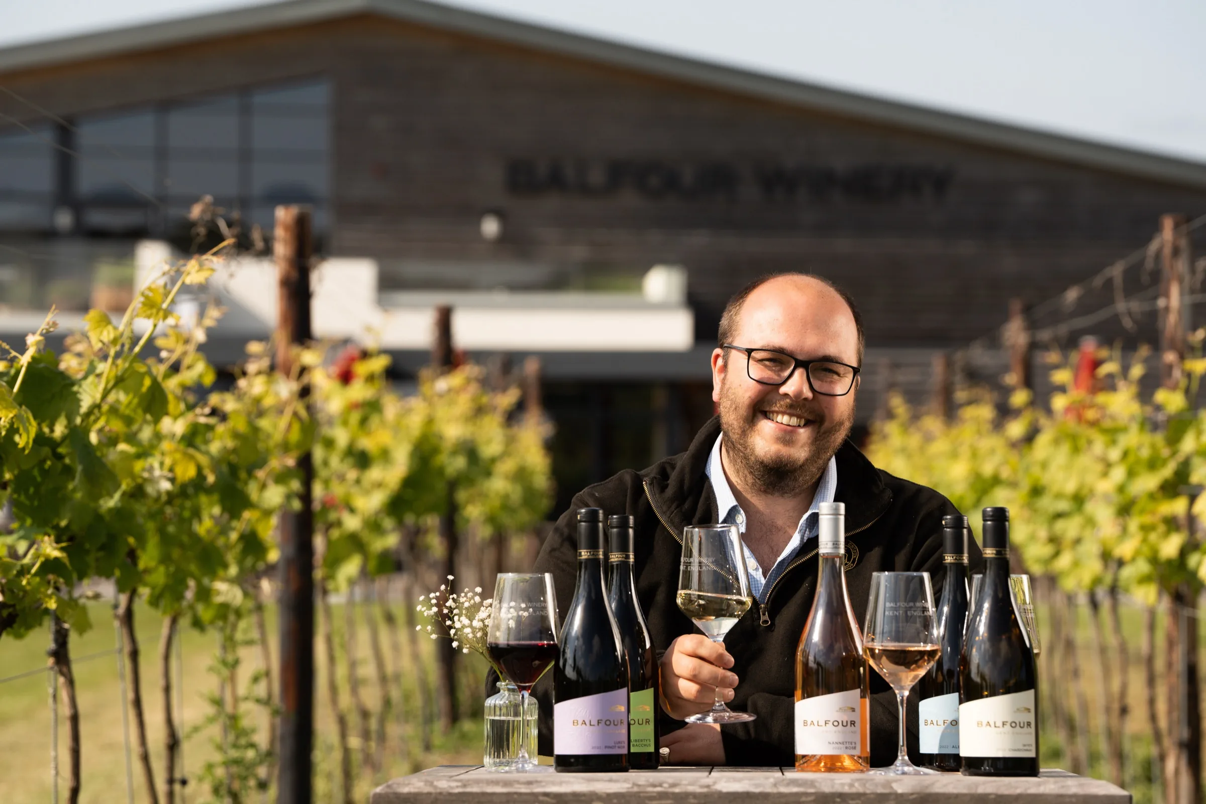A smiling man raising a glass of white wine among Balfour bottles in the estate vineyard